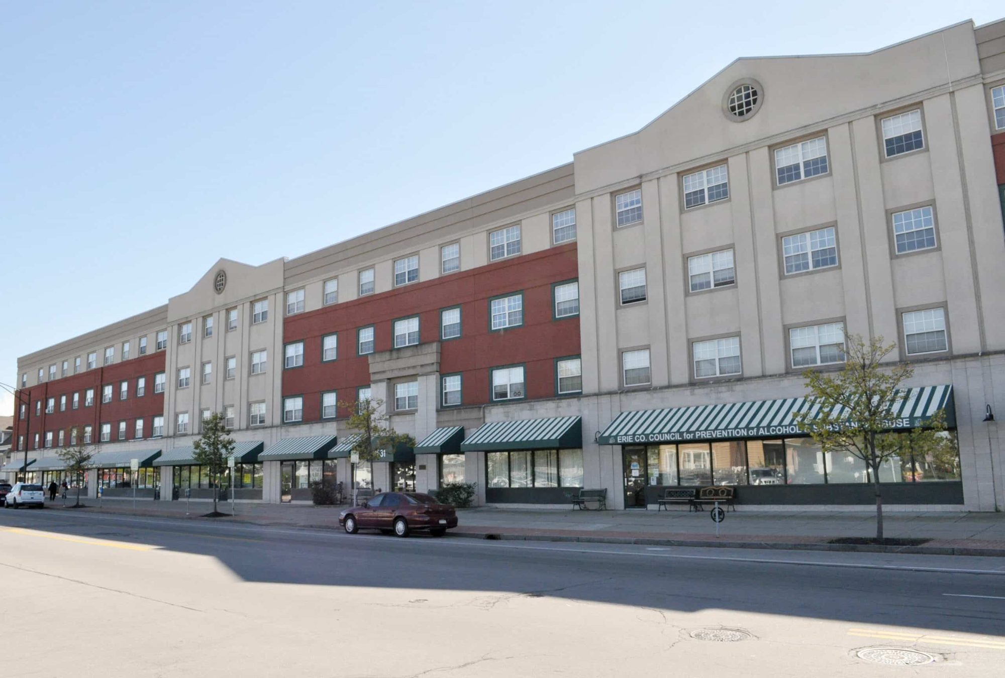 Hertel Park exterior — four-story mixed-use building with red brick and cream stucco façade and awning-shaded storefronts along Hertel Avenue