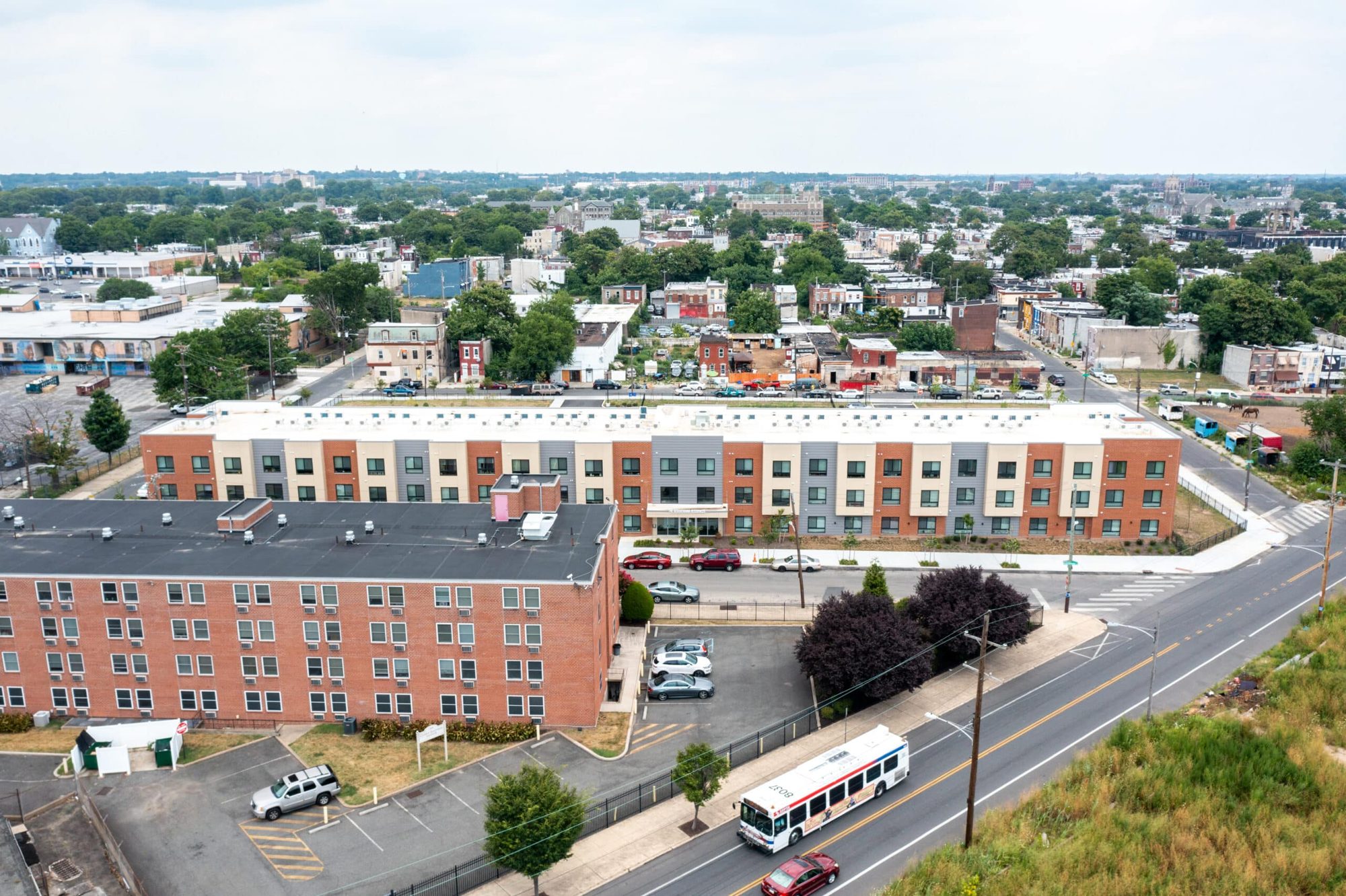 Susquehanna aerial view — three-story brick, cream, and gray-paneled affordable apartment building along a North Philadelphia corridor with the city skyline beyond